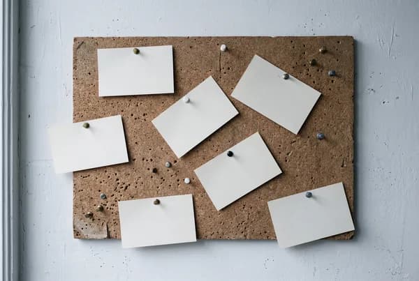 A cork noticeboard on a white wall with blank index cards pinned at angles and several empty pins where cards have been taken.