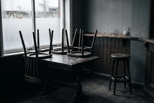 An empty pub corner after closing with chairs upturned on a dark table and one stool still standing on the floor beside it.