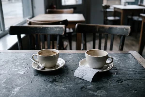 Two empty coffee cups on a cafe table with a crumpled receipt under one saucer and both chairs turned outward.