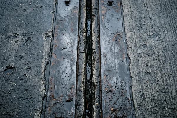 A concrete bridge expansion joint seen straight down, the gap running through the frame between weathered steel plates wet from rain.